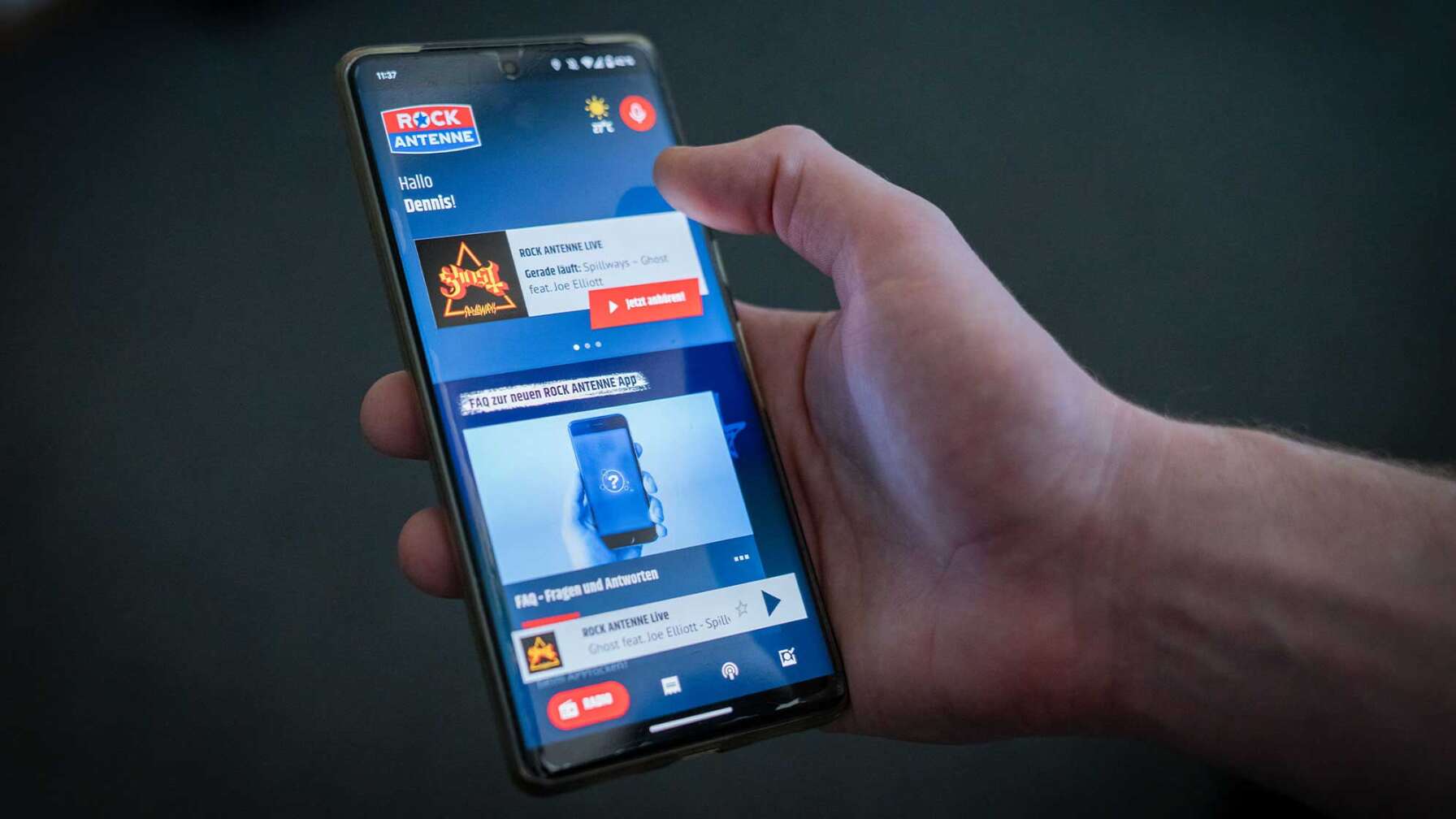 Eine Hand, die ein Smartphone in der Hand hält, auf dem die ROCK ANTENNE App geöffnet ist.