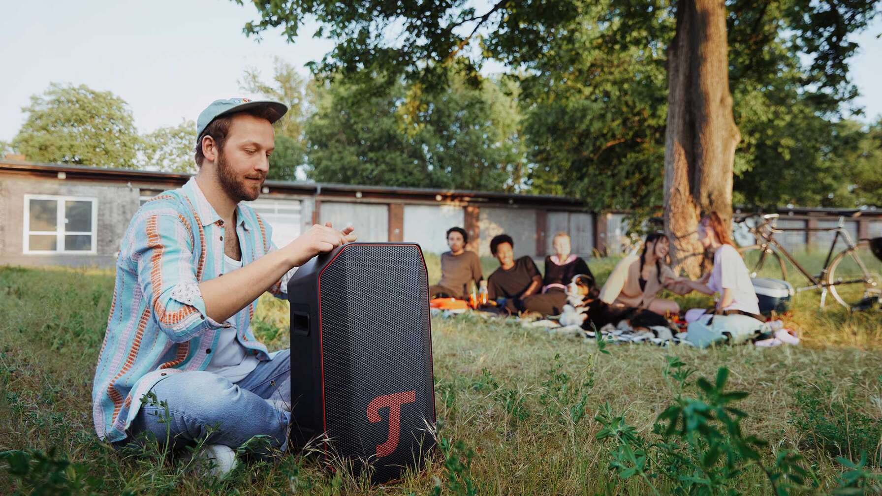Ein junger Mann, der auf einer Wiese sitzt und einen großen Lautsprecher der Marke Teufel bedient.