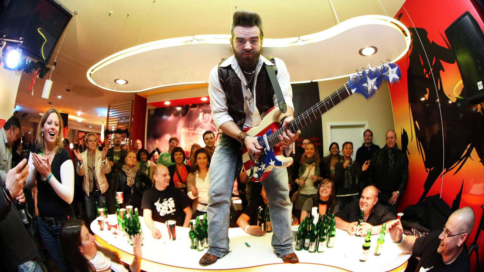 Bild eines Musikers mit Gitarre, der in einer Bar voller jubelnder Menschen auf dem Tresen steht und spielt